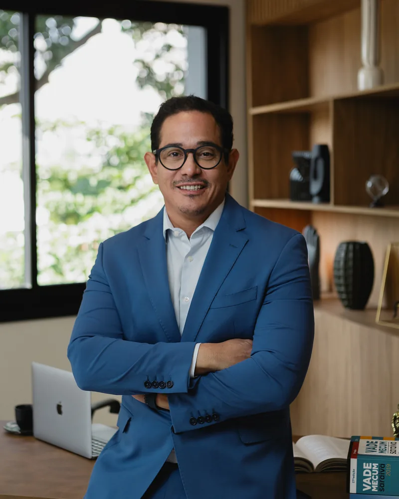 Dr. Daniel Oliveira sorrindo em seu escritório, de braços cruzados.