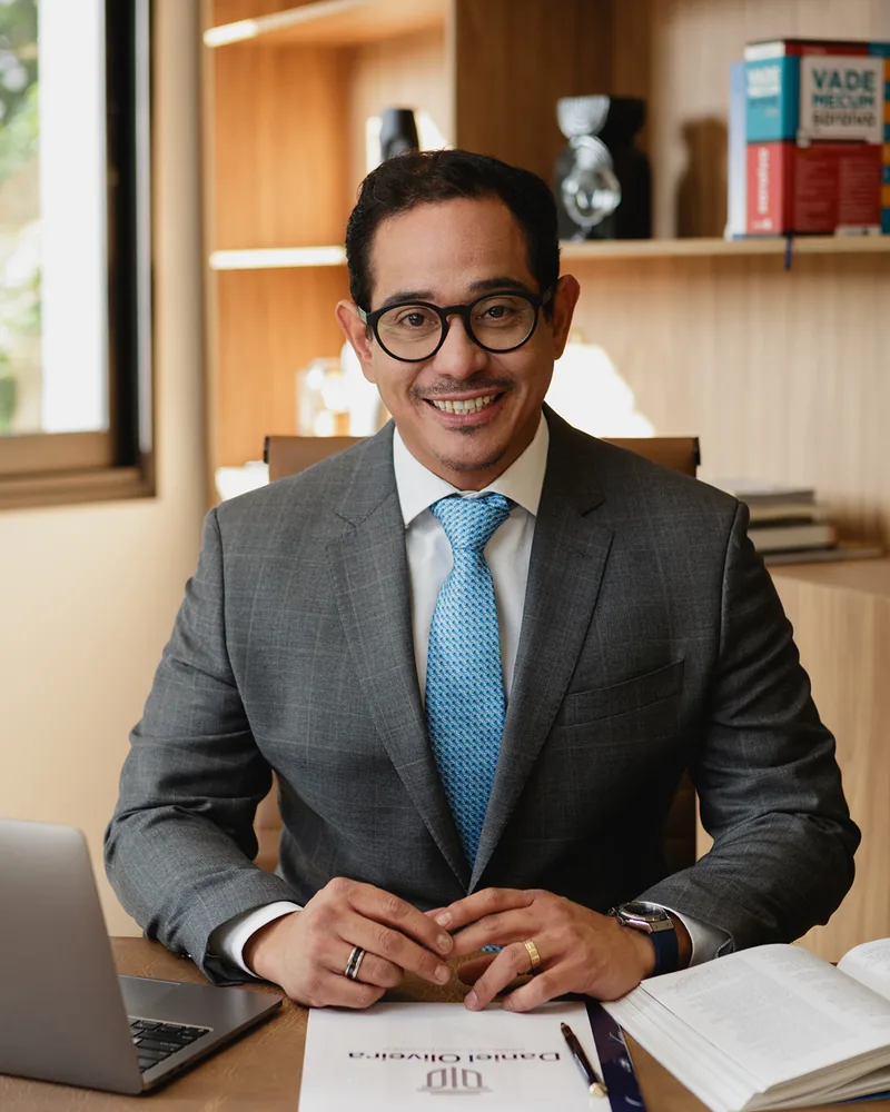 Dr. Daniel Oliveira sorrindo à mesa de trabalho, com livros e estante ao fundo.